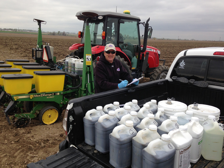 A man with a clipboard standing in front of a tractor and behind a truck bed filled with plastic 2.5 gallon jugs